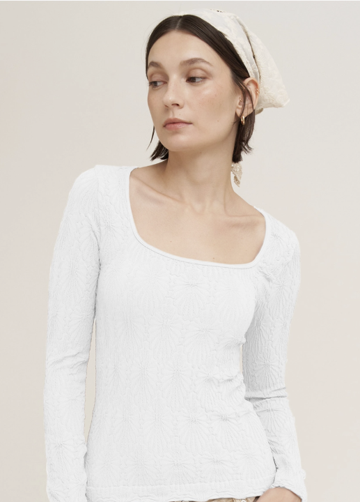A woman wearing a white textured long-sleeve top with a wide scoop neckline, paired with a cream-colored headscarf, photographed against a solid light beige background.