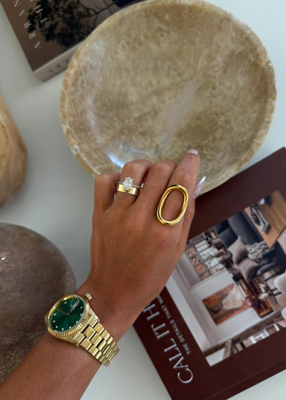 Hand wearing a gold ring and watch, holding a textured bowl with books in the background