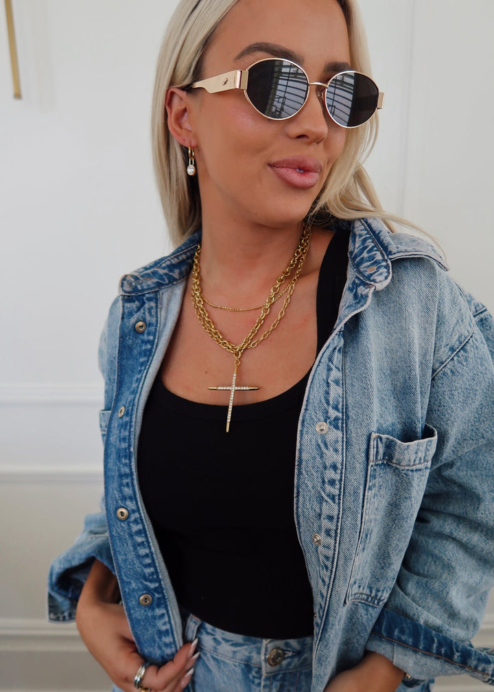 A woman wearing round gold-framed sunglasses, layered gold necklaces including a cross pendant, a black tank top, and a light-wash denim jacket over light-wash denim jeans.