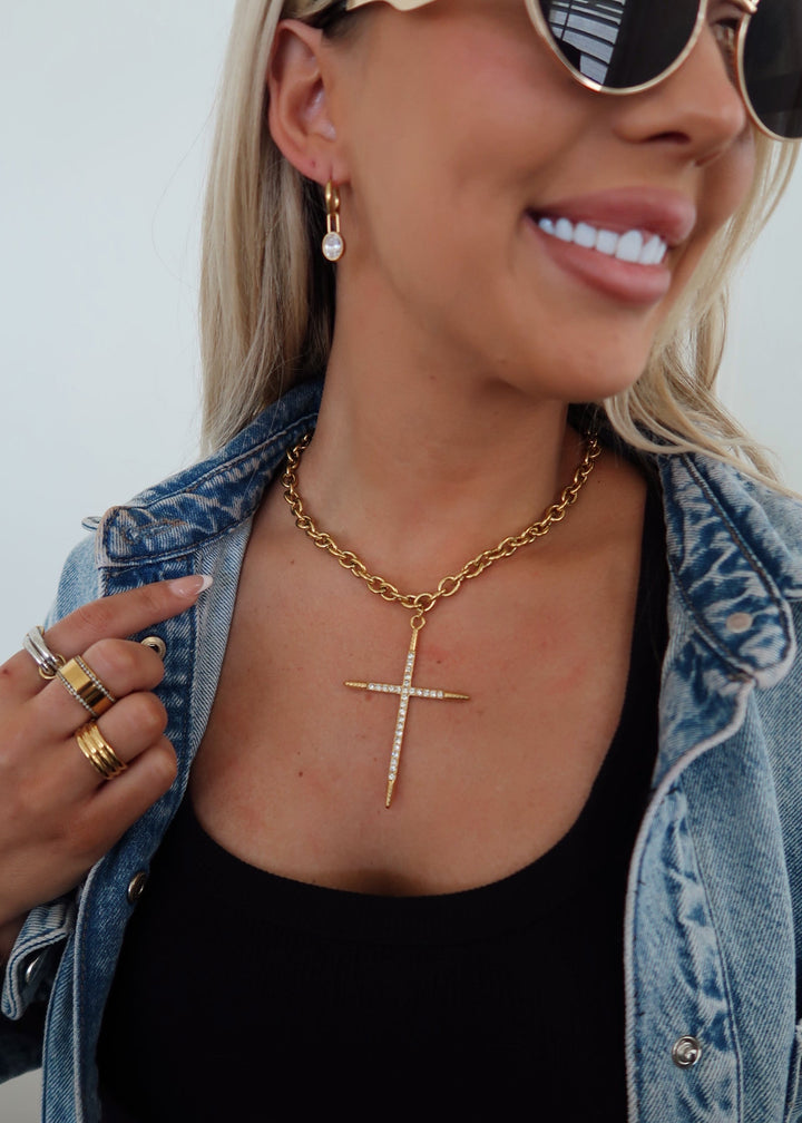 Woman wearing a gold necklace with a cross pendant, denim jacket, and sunglasses.