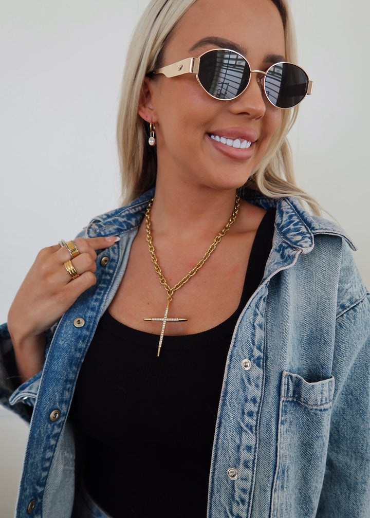 Woman wearing sunglasses, a denim jacket, and black top with gold jewelry.