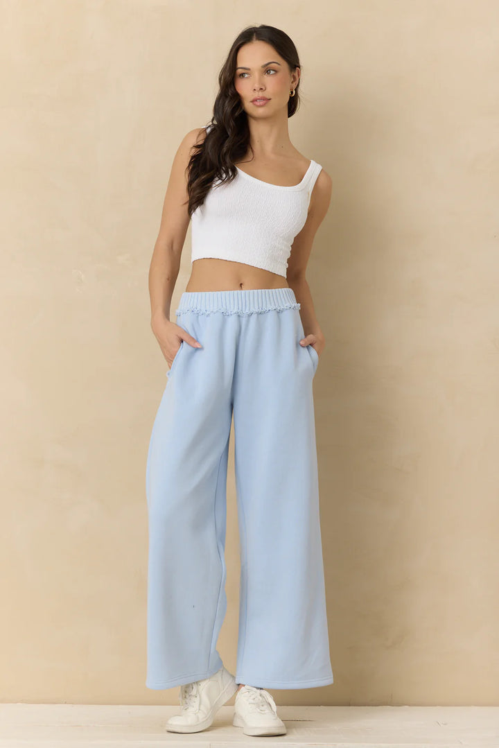 Woman wearing a white tank top and light blue pants against a beige background
