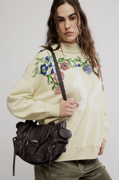 Woman wearing a cream-colored sweatshirt with floral embroidery, holding a black bag against a plain background