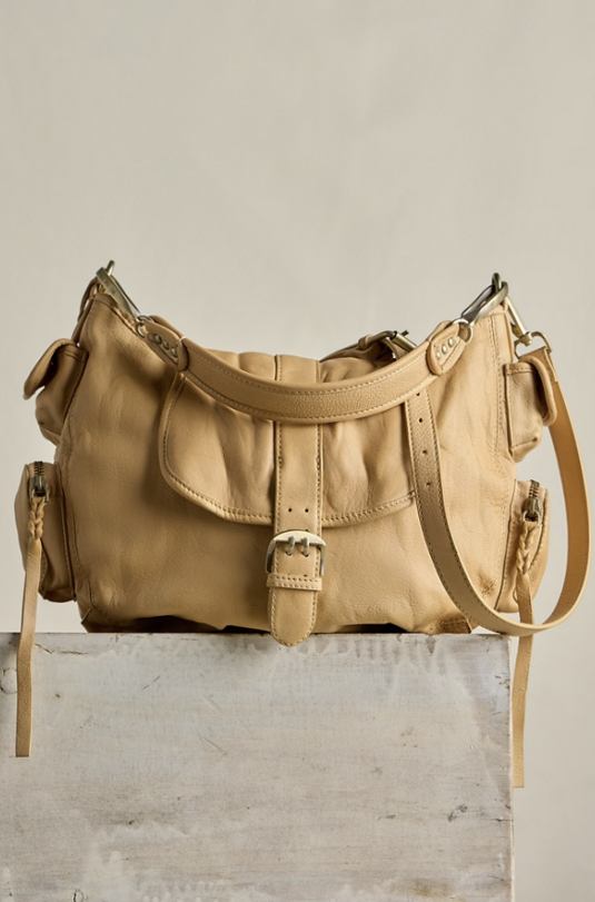 A tan leather shoulder bag with a flap front, buckle detail, and side pockets, displayed on a rustic wooden block against a neutral beige background.
