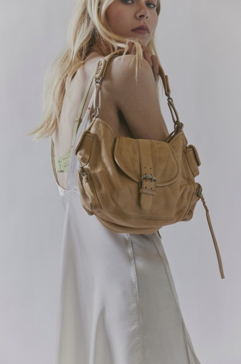 A woman wearing a white satin dress carries a tan leather shoulder bag over her shoulder against a light gray studio background.