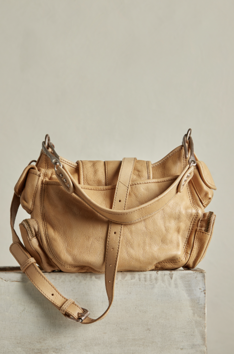 A tan leather shoulder bag with a pebbled texture, displayed on a light gray stone pedestal against a neutral beige background.