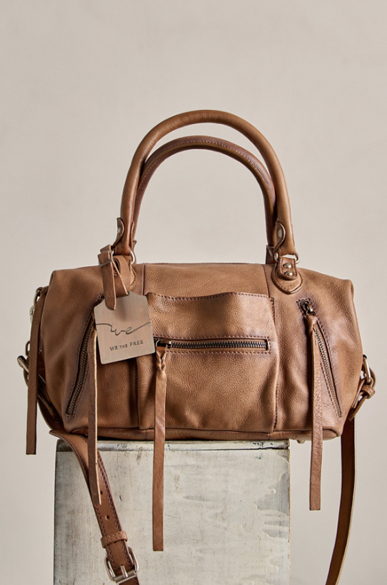 A tan leather handbag with double top handles, a front zip pocket, and a hanging brand tag, displayed on a textured pedestal against a neutral beige background.
