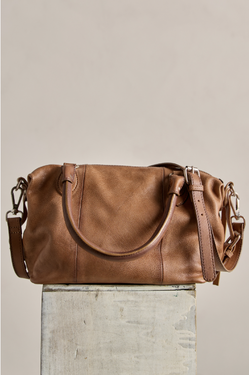 A medium brown leather duffel bag with a slouchy shape, double top handles, and a detachable shoulder strap, displayed on a textured gray block against a solid beige background.