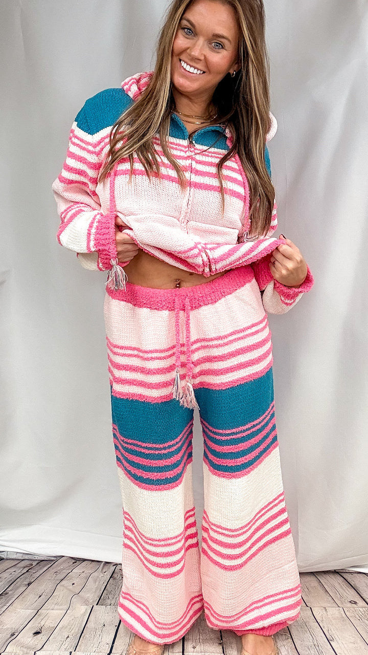 Woman wearing matching stripe top and bottom with pink, teal and cream horizontal stripes on plain background.