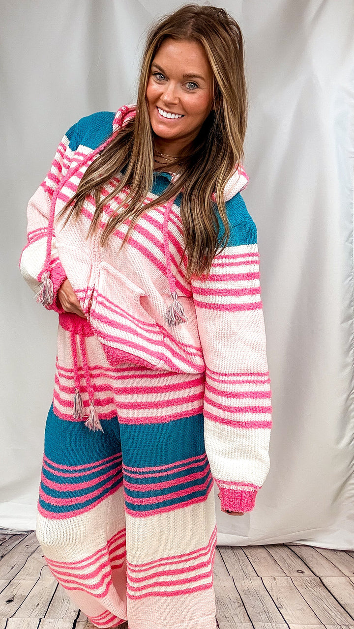 A woman modeling a matching knit lounge set consisting of a hooded sweatshirt and wide-leg pants, featuring a color-blocked pattern of pink, teal blue, and cream stripes.