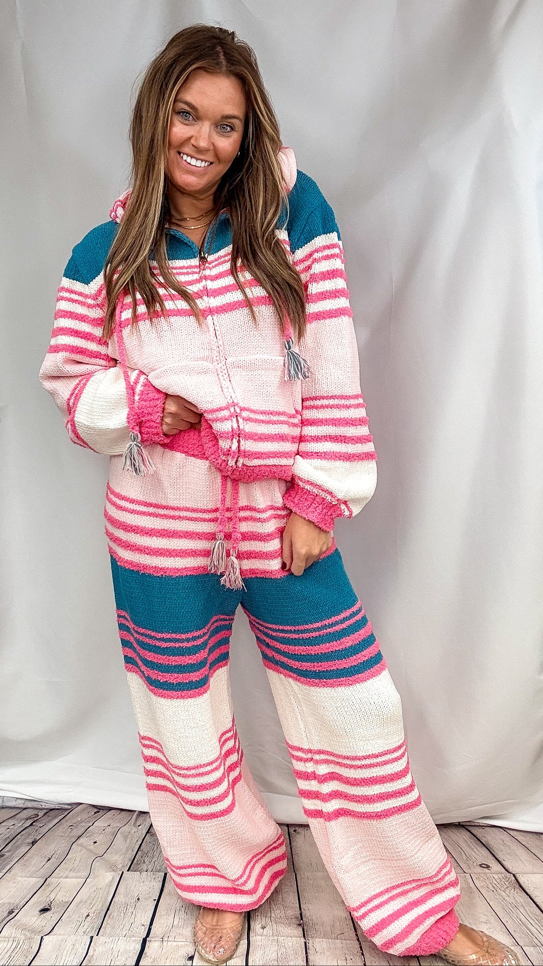 A woman modeling a matching knit lounge set with pink, teal, and cream horizontal stripes, standing against a white draped backdrop on a light wood floor.