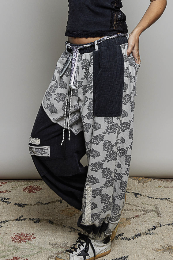 A person wearing loose, baggy patchwork pants with a gray and black floral print on one leg and solid black fabric on the other, styled with a black top and sneakers against a plain wall and patterned rug.