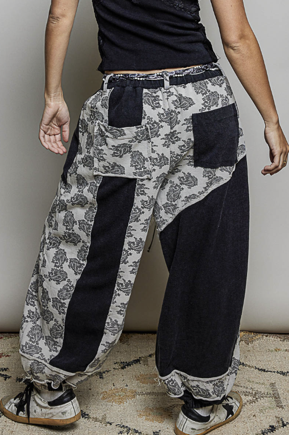 A person shown from the back wearing loose, patchwork-style pants with a black-and-white floral print and solid black side panels, paired with a black top and sneakers against a plain indoor wall.