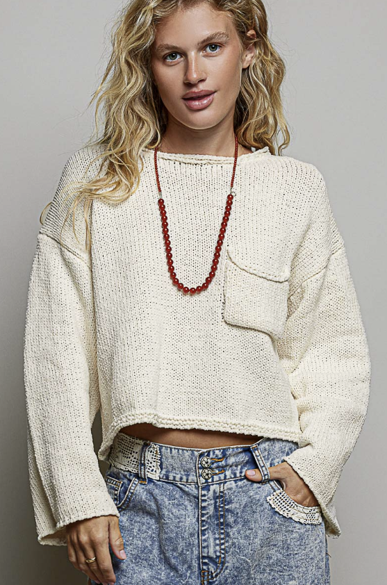 A woman wearing a cream-colored, textured knit long-sleeve crop top sweater with a chest pocket, paired with blue denim jeans and a red beaded necklace, against a solid gray studio background.