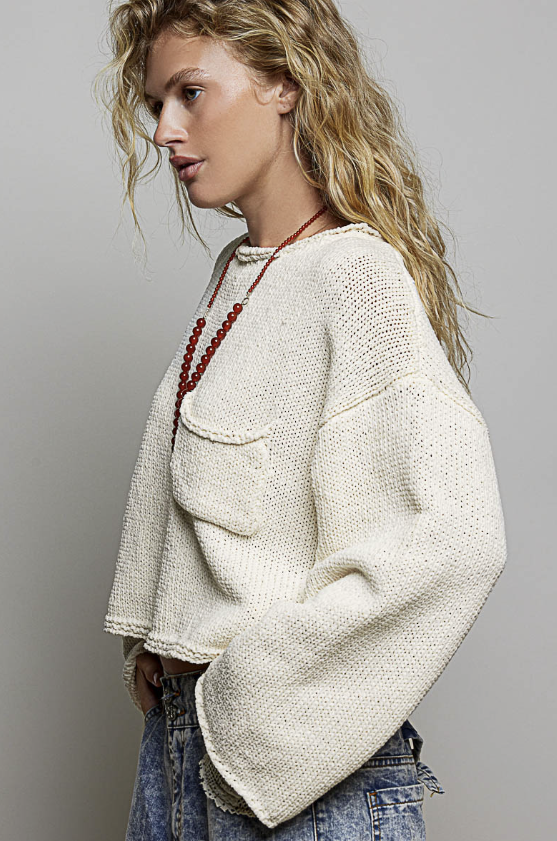 A woman wearing a cream-colored, textured knit sweater with a chest pocket, paired with blue jeans and a red beaded necklace, shown in a side profile against a neutral gray background.