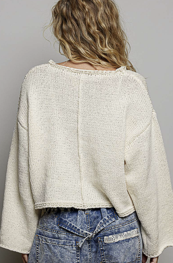 A woman viewed from behind wearing a cream-colored, textured knit cropped sweater and blue jeans against a plain gray studio background.