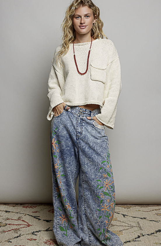 A woman stands against a plain light gray wall wearing a cream knit cropped sweater, wide-leg blue jeans with orange and green floral embroidery, and a long red beaded necklace.