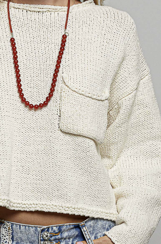 Close-up of a person wearing a chunky knit off-white cropped sweater with a chest pocket, paired with a long red beaded necklace and blue jeans.