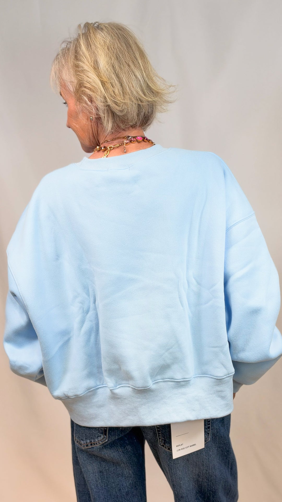 Woman shown from back wearing light blue sweatshirt and jeans on plain background