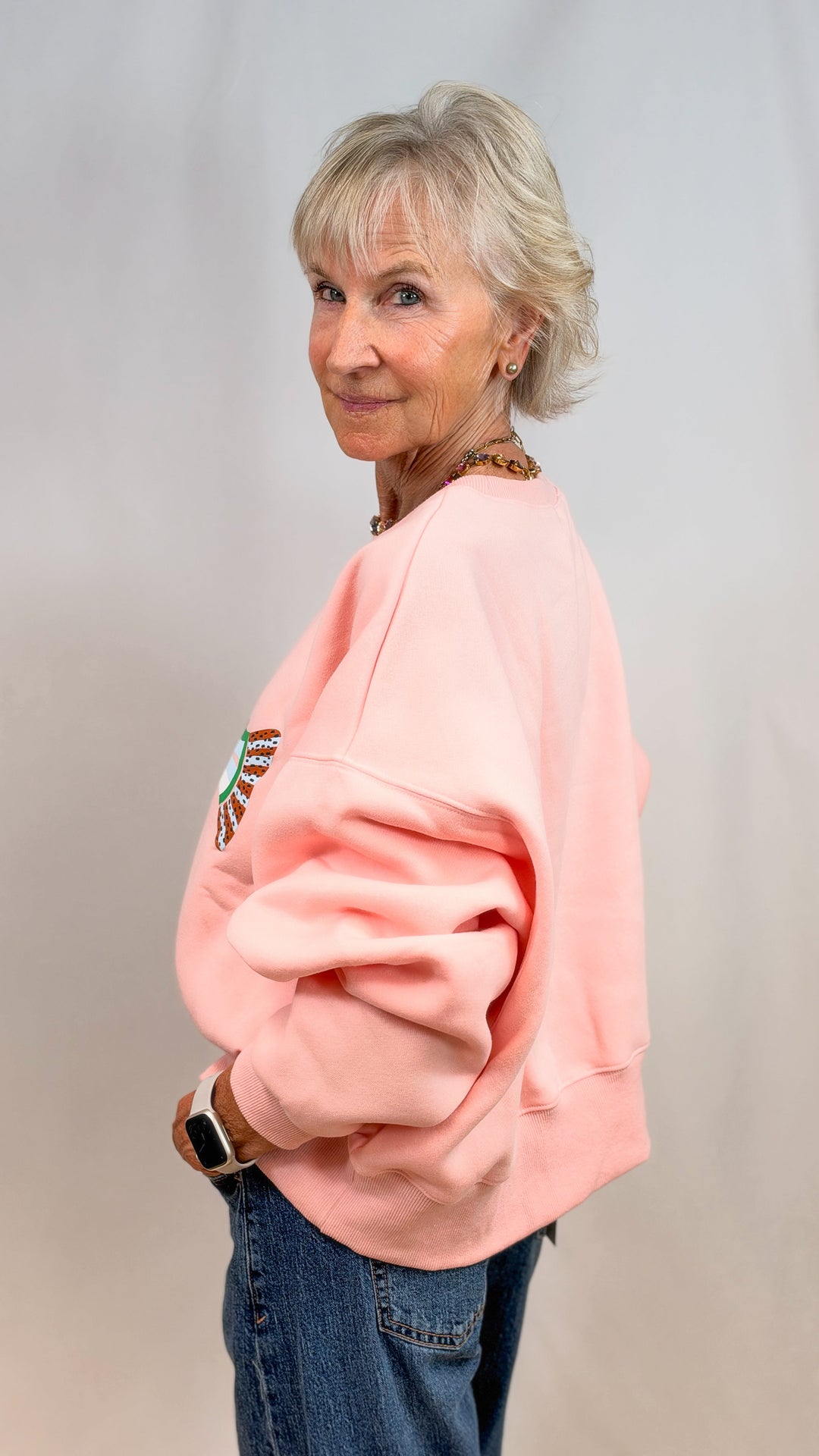 Woman shown from side view wearing salmon sweatshirt and jeans on plain background