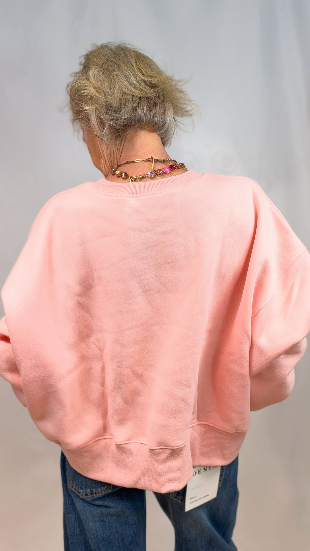 Woman shown from back wearing salmon color sweatshirt and jeans on plain background