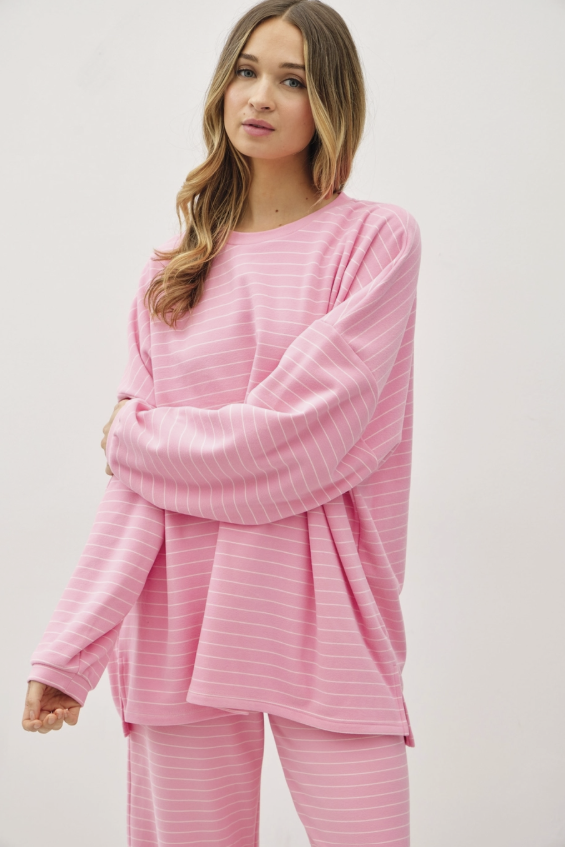 A woman modeling a pink two-piece pajama set with thin white horizontal stripes, featuring a long-sleeve top and matching pants, against a plain light gray studio background.