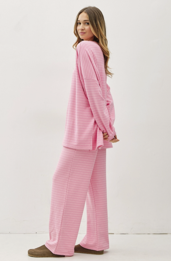 A woman modeling a pink long-sleeve pajama set with thin white horizontal stripes, consisting of a loose long-sleeve top and matching wide-leg pants, paired with brown slippers against a plain white studio background.