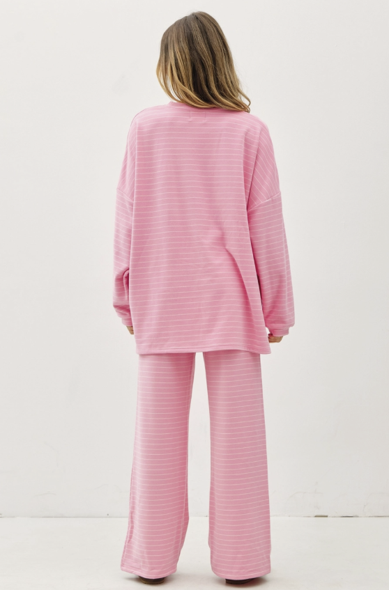 A woman viewed from behind wearing a matching pink two-piece lounge set with thin white horizontal stripes, consisting of a long-sleeve top and wide-leg pants, against a plain white studio background.