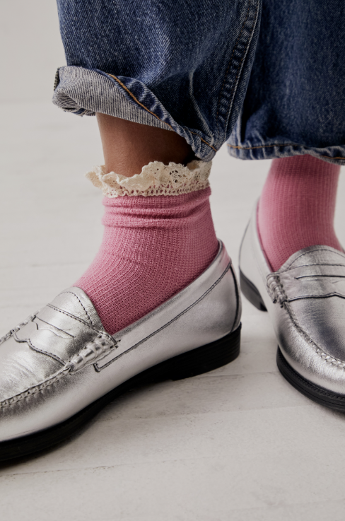 Pink ribbed crew socks with a white lace ruffle trim, worn with silver metallic penny loafers and cuffed blue jeans.
