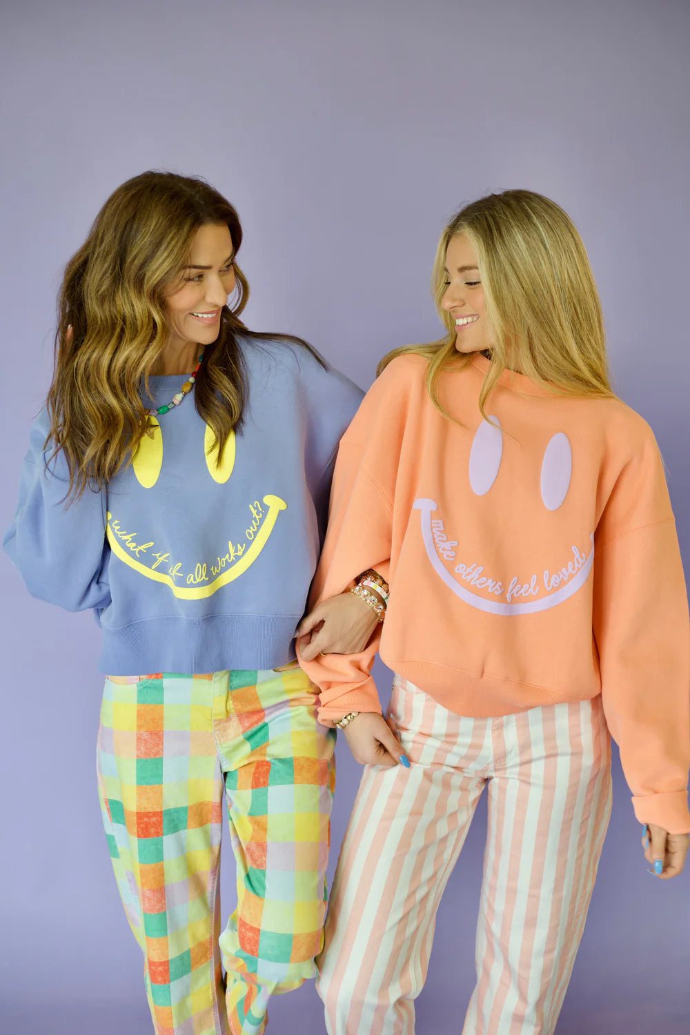 Two women wearing colorful sweaters and pants against a purple background