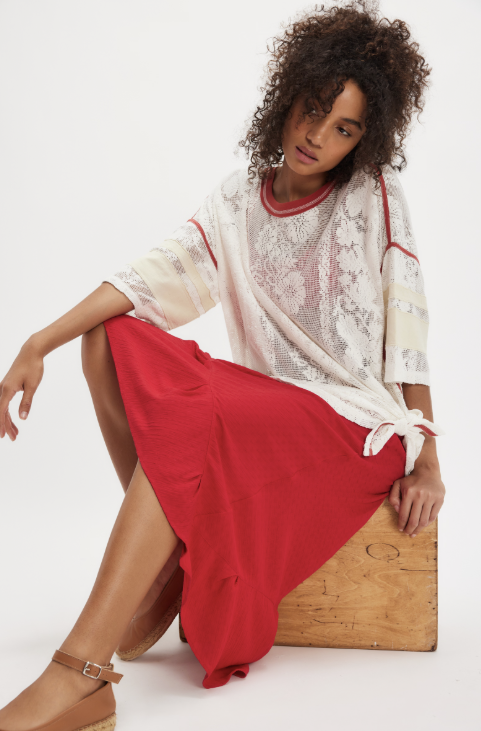 Woman wearing a white lace top and red skirt sitting on a wooden box against a white background