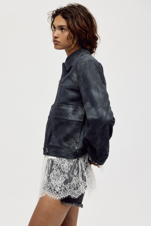 A model shown in profile wearing a dark washed leather jacket over a white lace hem shirt and dark denim shorts, photographed against a solid light gray studio background.