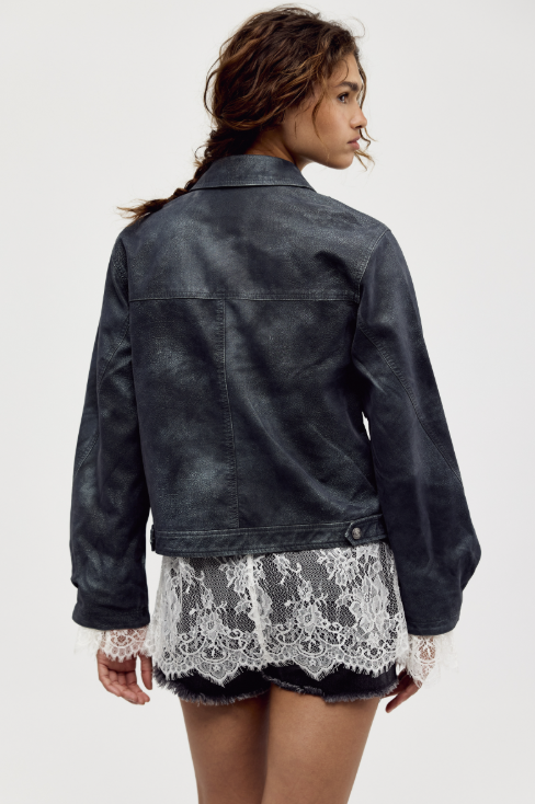 A woman shown from the back wearing a dark washed leather jacket over a white lace mini skirt, against a plain light gray studio background.