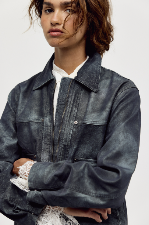 A close-up of a model wearing a dark gray, washed denim-style jacket over a white collared shirt and white lace cuffs, set against a plain light gray background.
