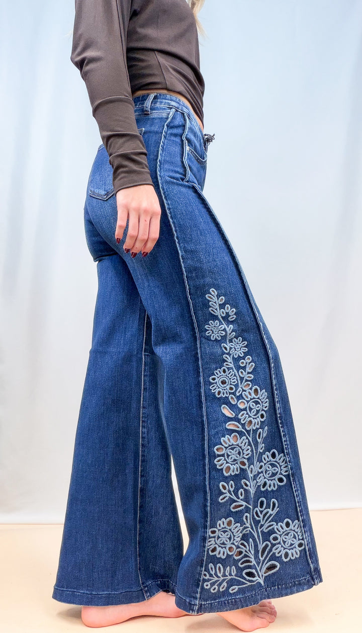 Woman wearing medium wash denim wide leg jeans with cutout floral pattern on the sides on plain background