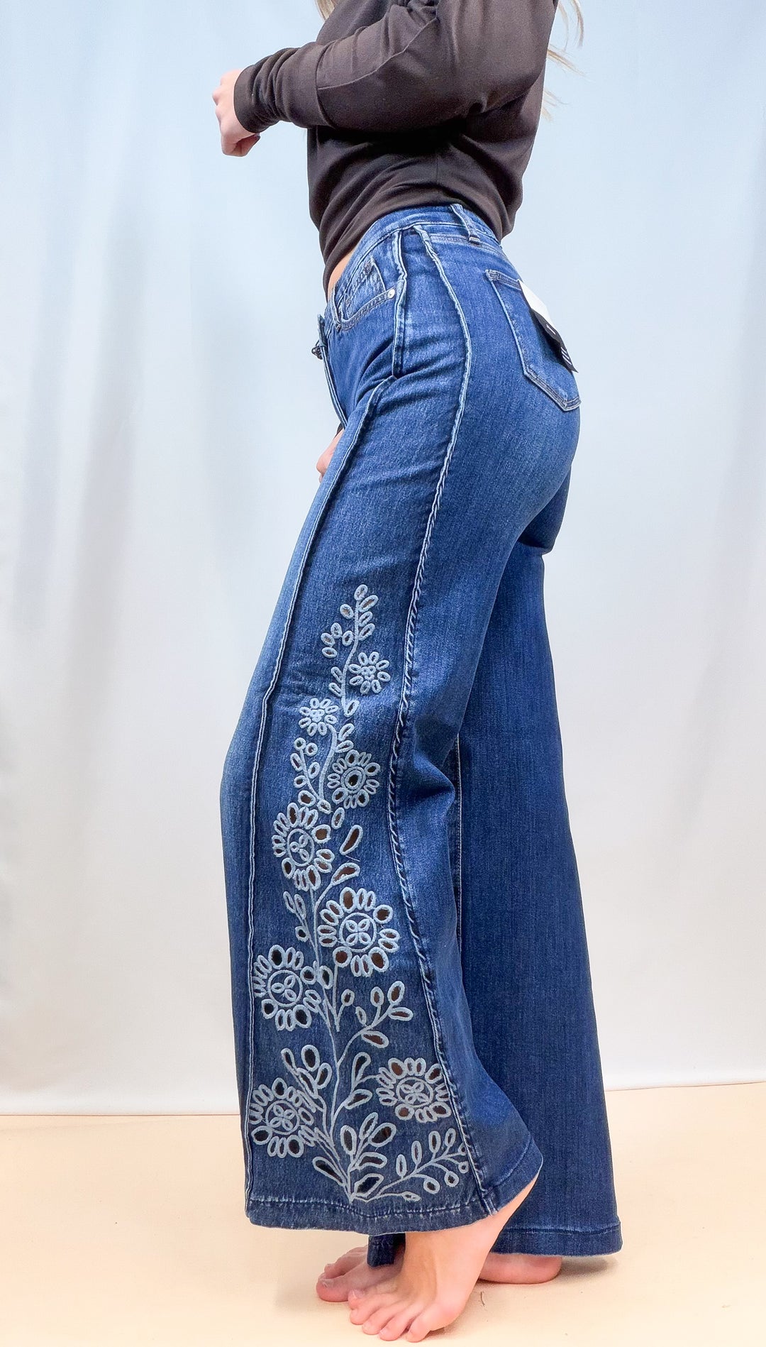 Woman shown from side view wearing medium wash denim jeans with cutout floral on the sides on plain background