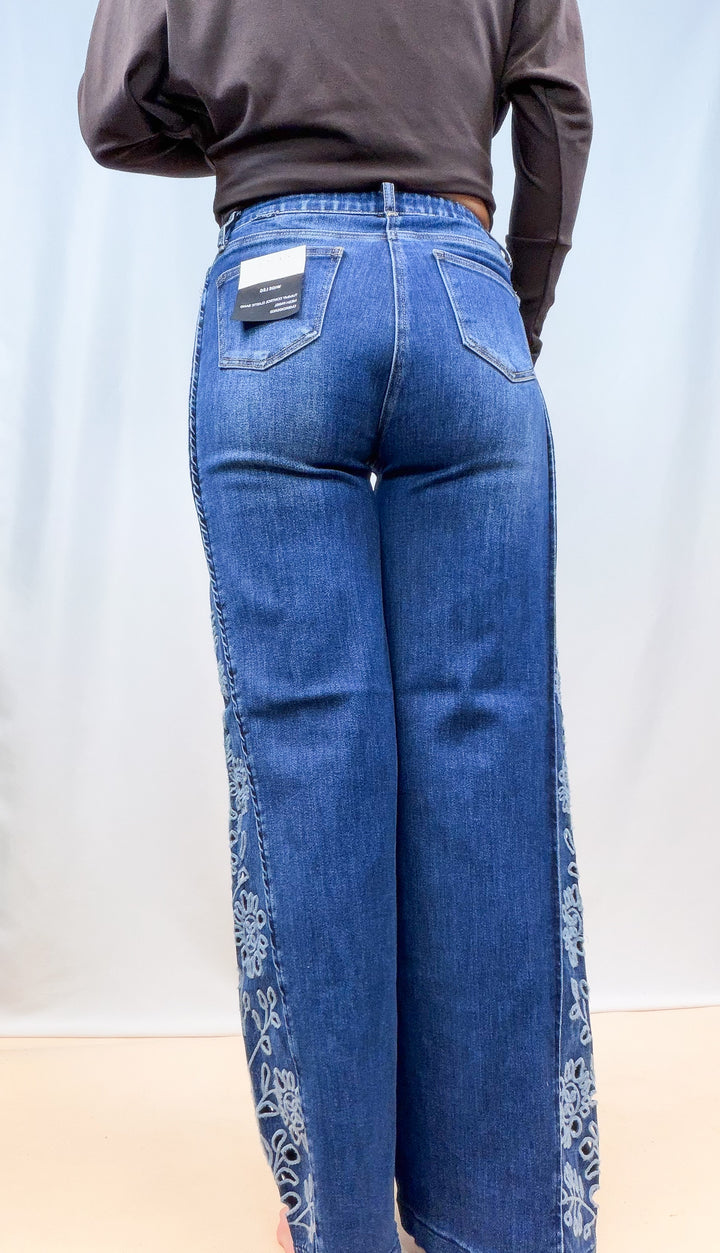 Woman shown from back view wearing medium wash denim jeans with floral cutouts on sides on plain background