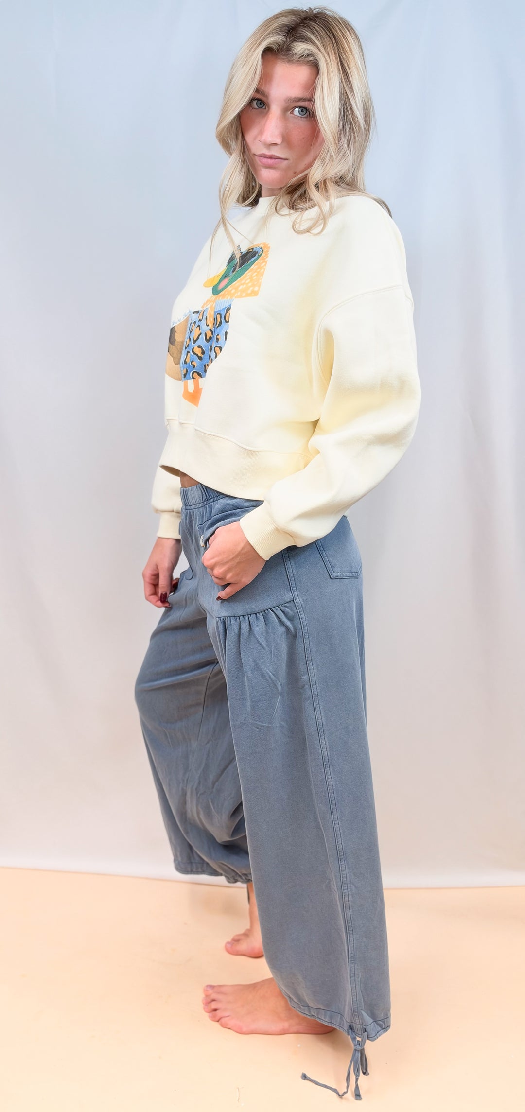 Woman shown from side view wearing duck graphic sweatshirt with blue cinch hem pants on plain background