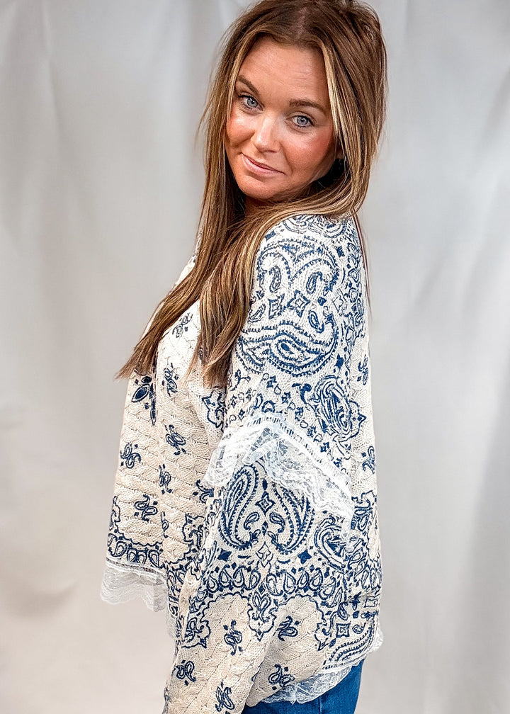 A woman wearing a cream-colored lace and paisley embroidered top with blue jeans, photographed against a white draped backdrop.