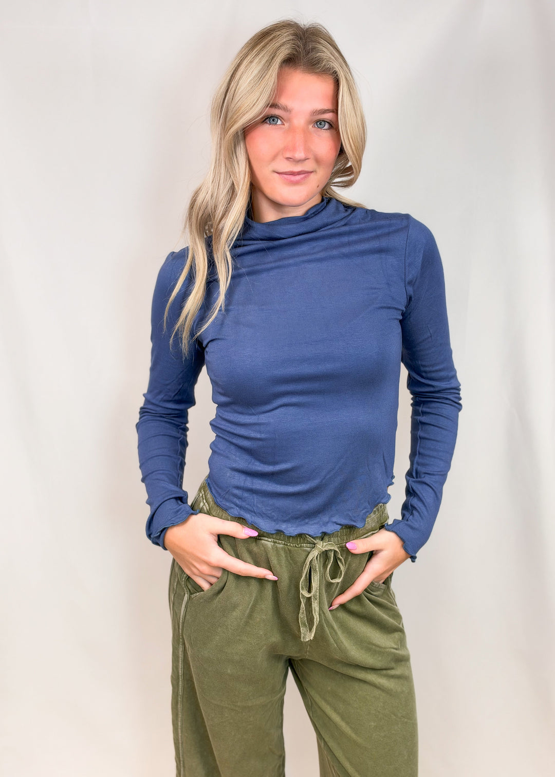 Woman wearing blue mock neck top with lettuce detail edges and green pants on plain background