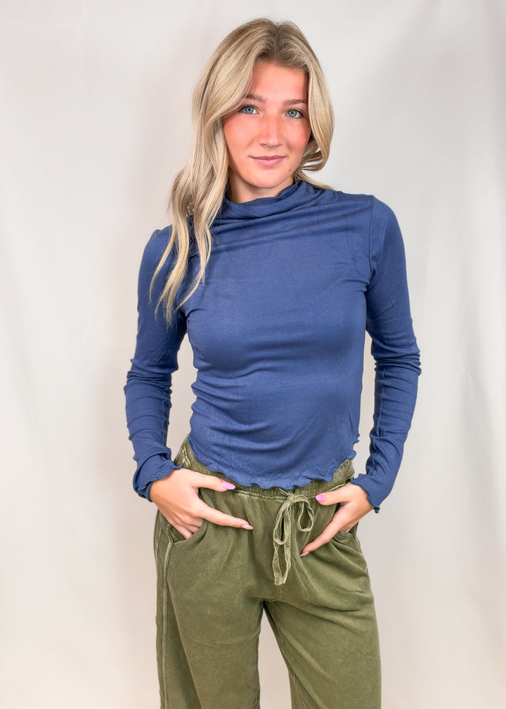 Woman wearing blue mock neck top with lettuce detail edges and green pants on plain background