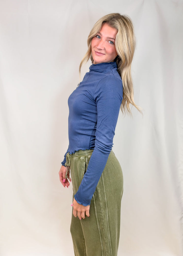 Woman wearing blue mock neck top with lettuce detail edges and green pants on plain background