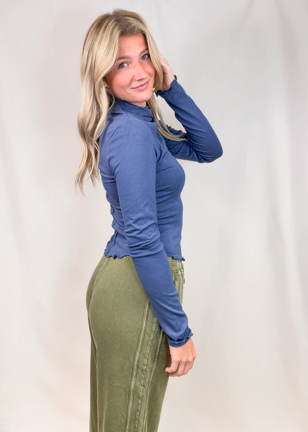 Woman shown from side view wearing blue mock neck top with lettuce detail edges and green pants on plain background
