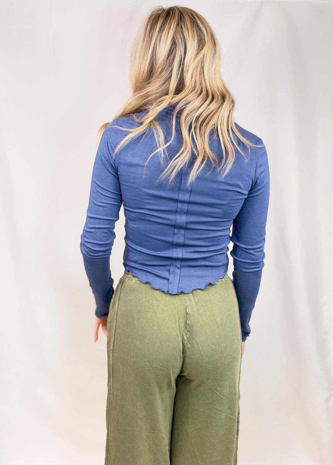 Woman shown from back wearing blue top and green pants on plain background