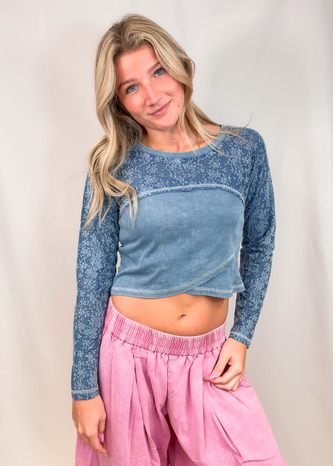 A woman modeling a blue long-sleeve crop top with a textured pattern on the sleeves and a solid blue front panel, paired with pink pants, against a light neutral studio backdrop.