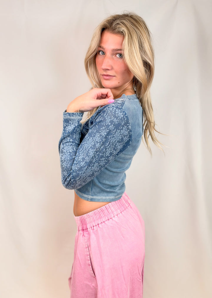 A blonde woman posing in a studio setting, wearing a blue long-sleeve textured crop top and pink drawstring sweatpants against a neutral beige backdrop.