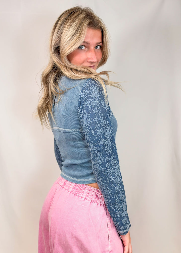 A woman with blonde hair is shown from the back wearing a cropped blue long-sleeve top with a textured floral pattern on one sleeve and a pink skirt, set against a plain light beige backdrop.