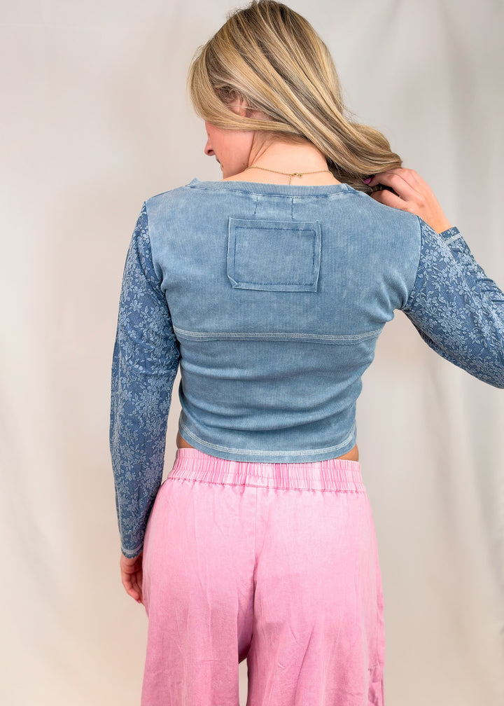 A woman shown from the back wearing a cropped long-sleeve top with a washed light blue body and mottled blue sleeves, paired with pink pants, against a plain light beige backdrop.