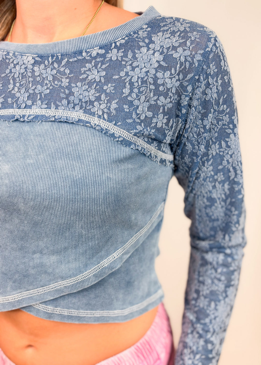 A close-up of a person wearing a cropped long-sleeve top with a light blue ribbed body and a sheer floral lace overlay across the shoulders and sleeves, paired with pink bottoms.