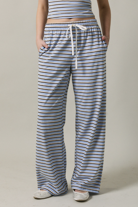 A person wearing loose, wide-leg striped pants with a white drawstring waist and side pockets, paired with a matching striped crop top and white shoes, against a plain beige studio background.